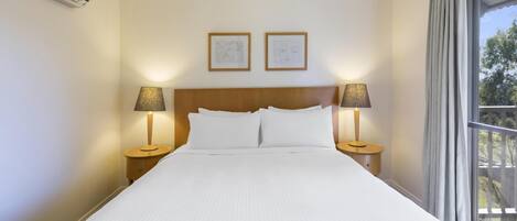 A hotel room with a large bed, two bedside lamps, an air conditioner, and two framed artworks on the wall.
