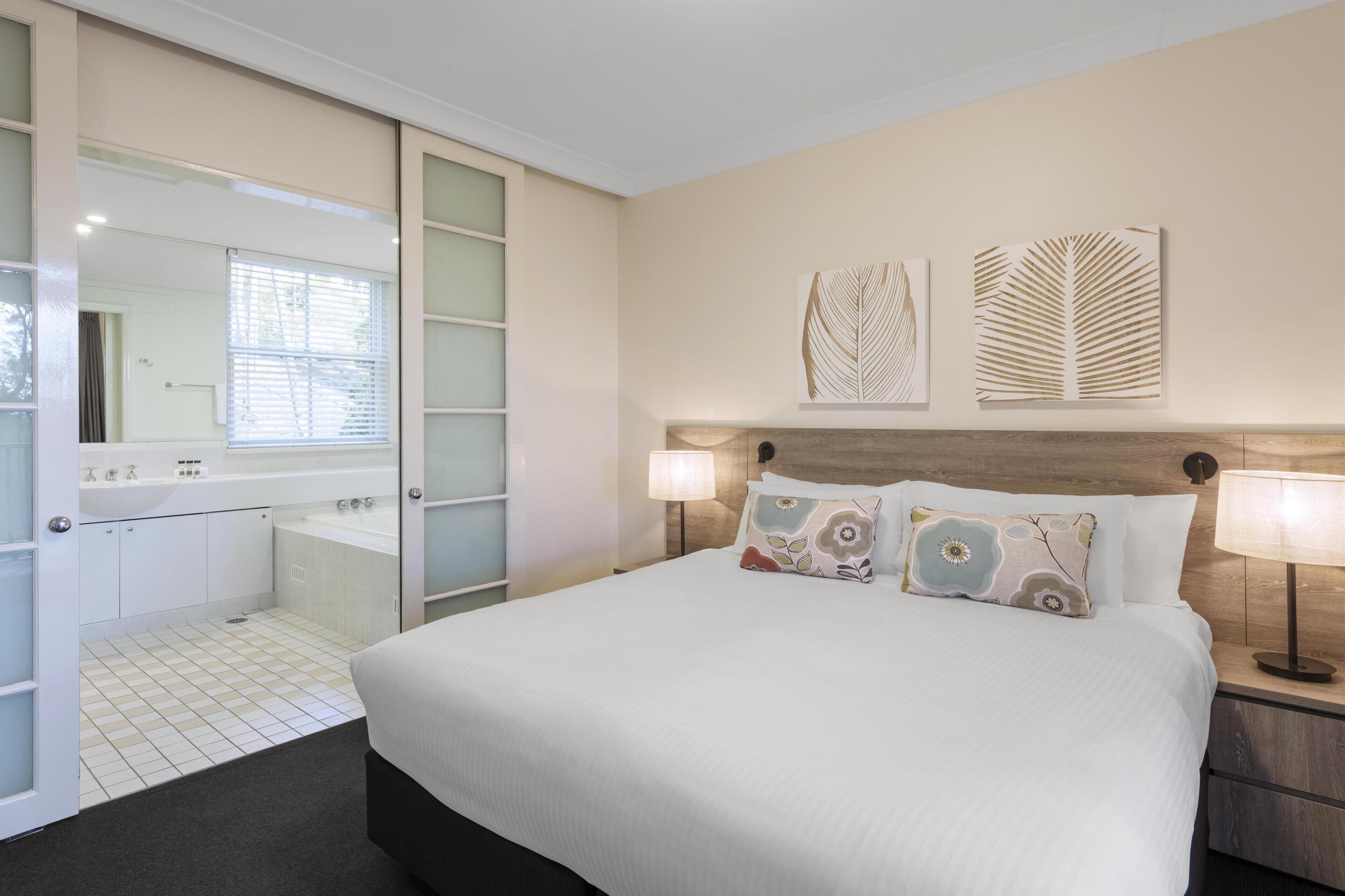 Habitación de hotel con una cama grande, dos lámparas de noche, una mesita de noche, un cabecero de madera y dos obras de arte en la pared.