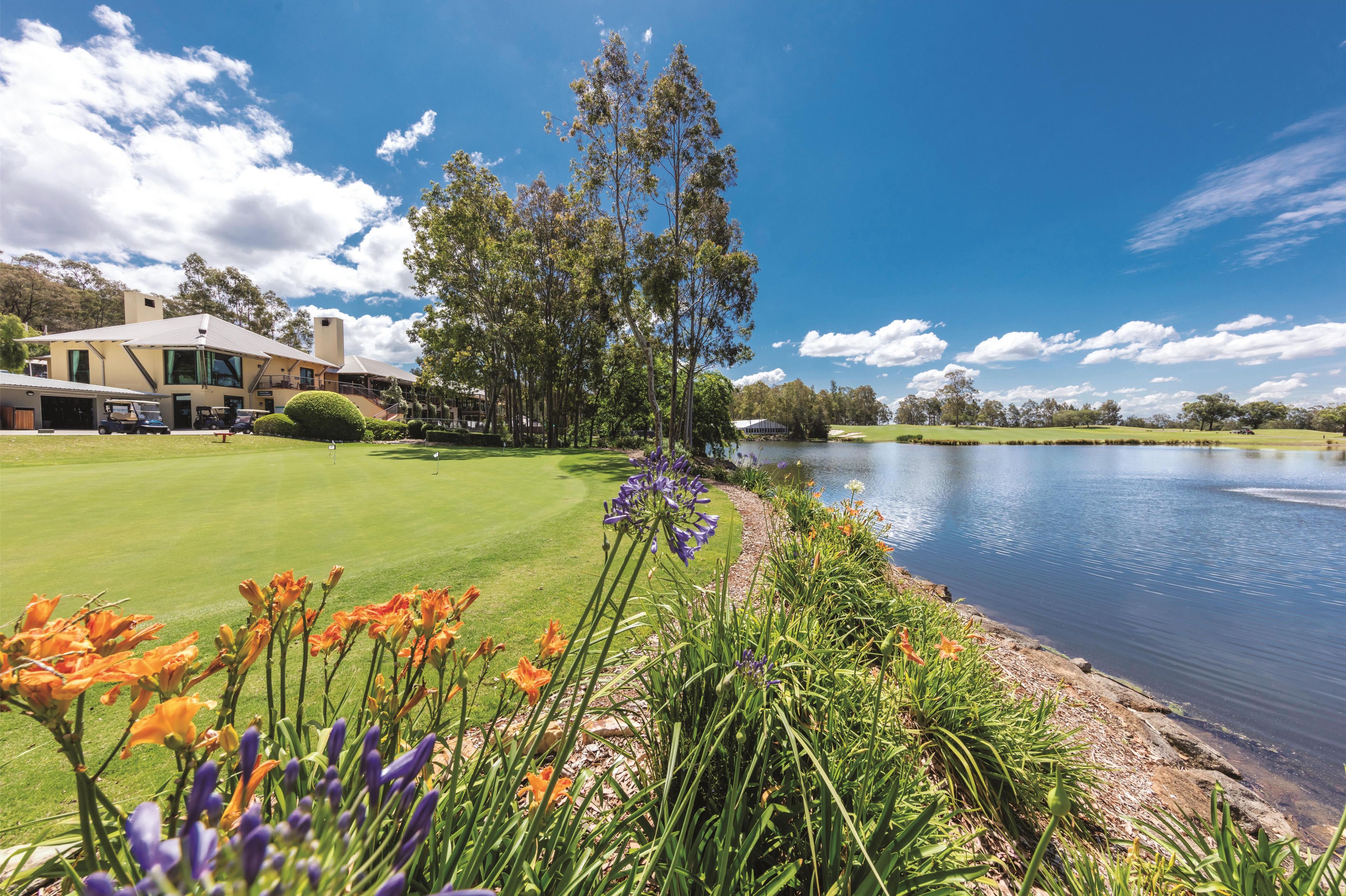 Un campo de golf con un club de campo, una masa de agua y diversas flores al frente.