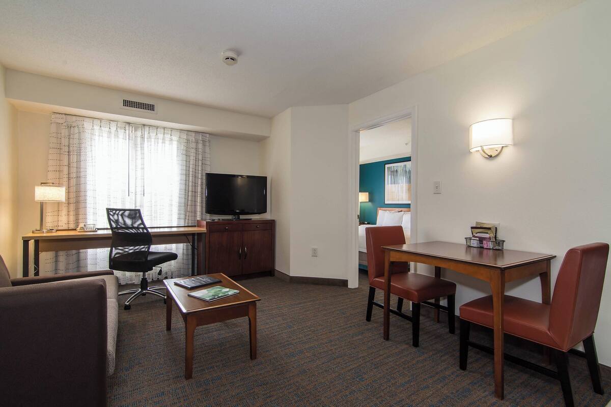 A hotel room with a desk, a television, a dining table with chairs, and a bed in the background.