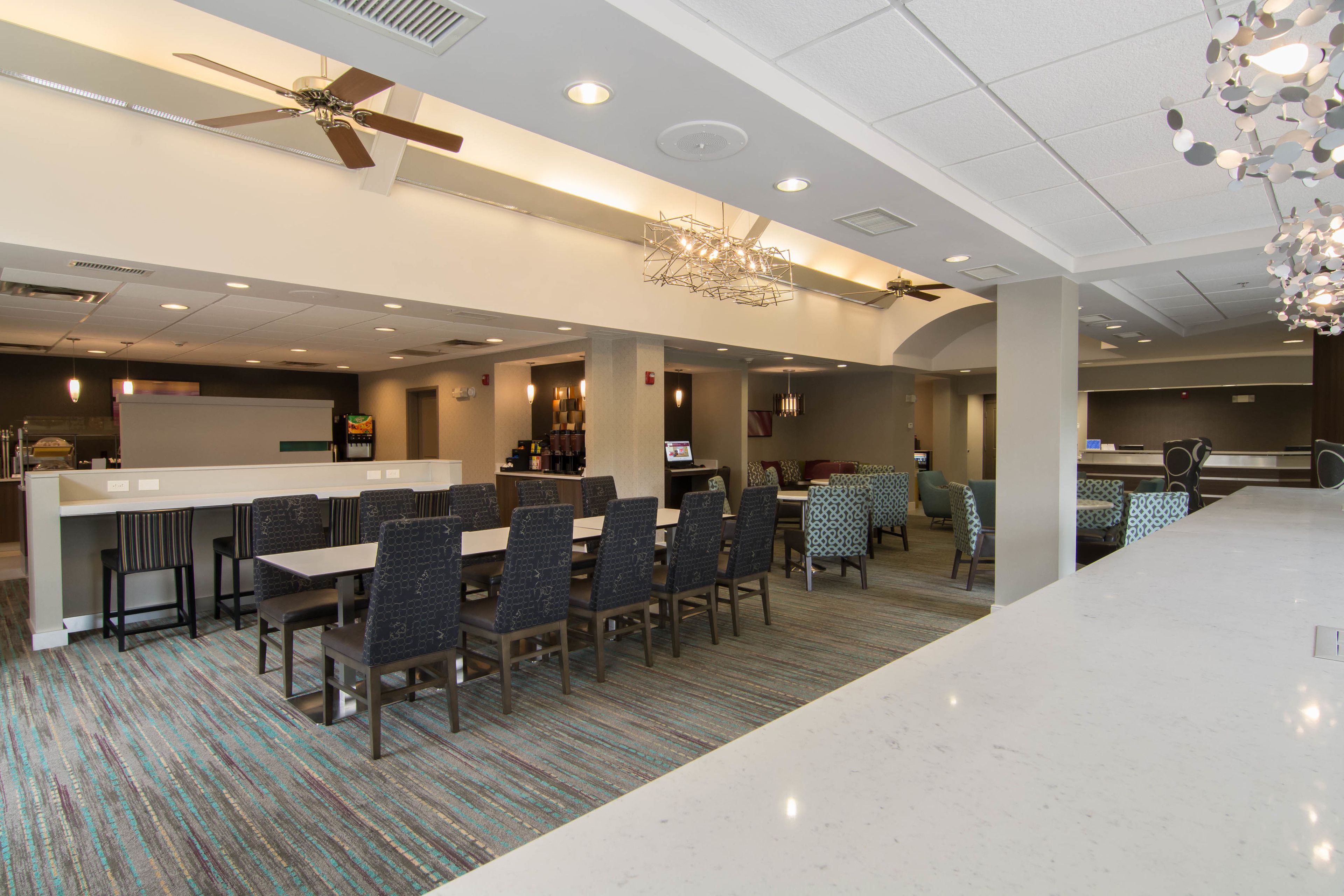 A spacious hotel lobby with a long reception desk, multiple seating areas, and a ceiling fan.