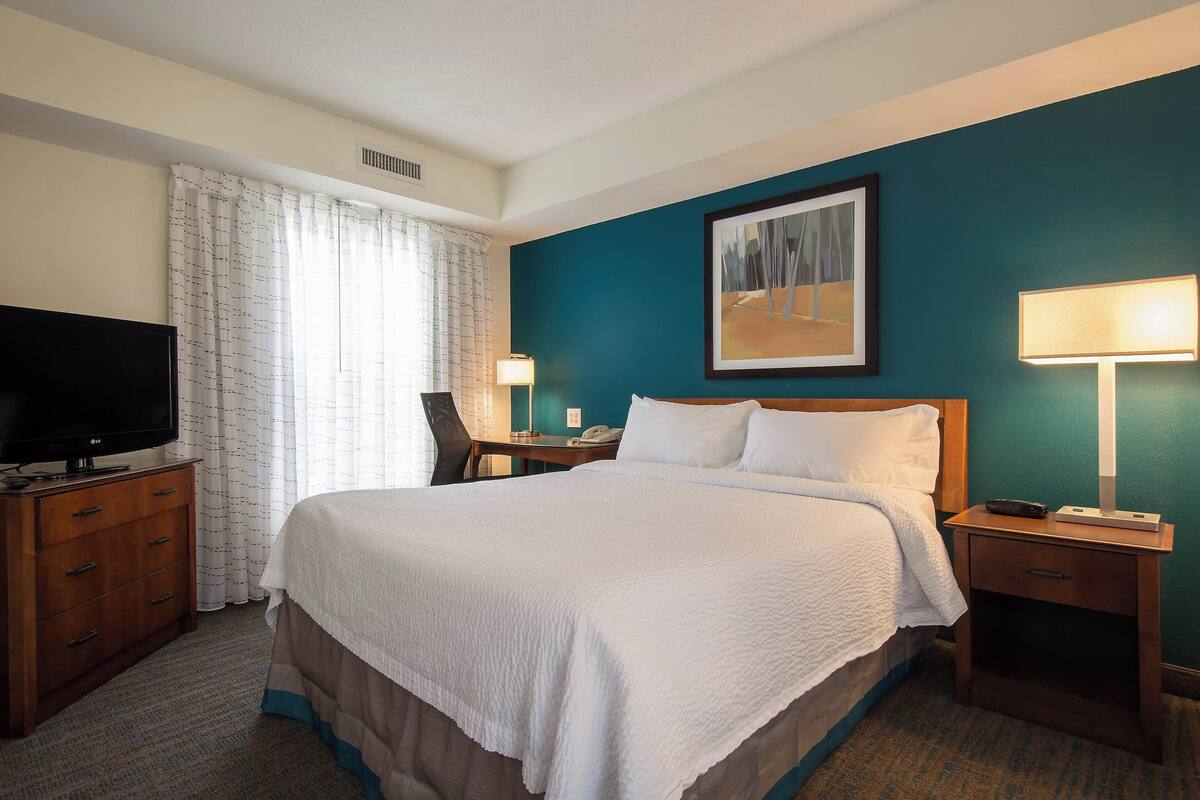 A hotel room with a large bed, a TV on a wooden dresser, a desk with a chair, and a painting on the wall.
