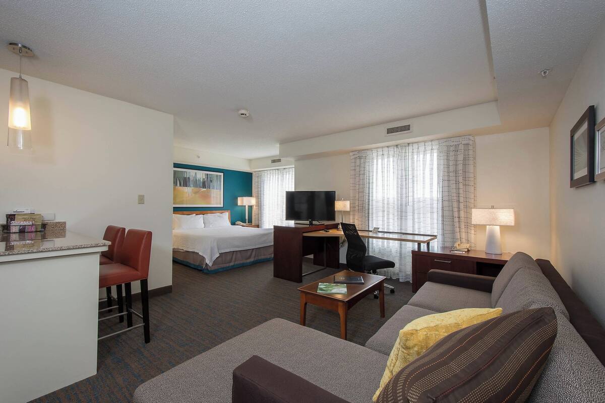 A hotel room with a bed, a desk with a computer, a TV, a sofa, and a small bar area.
