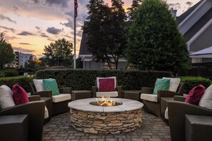 A stone fire pit surrounded by wicker furniture with cushions, set in a courtyard with a hedge and trees in the background. - Residence Inn by Marriott Charlotte Piper Glen (Charlotte)