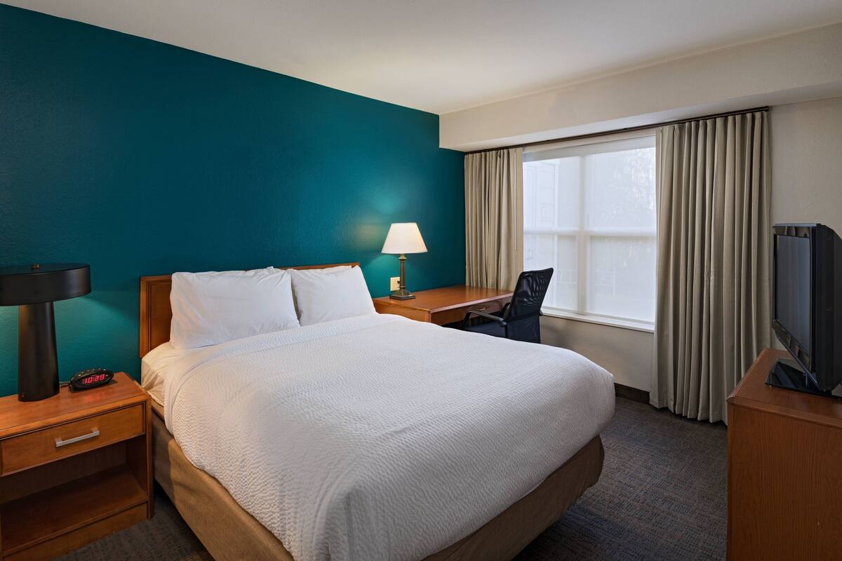 Una habitación de hotel con una cama grande, un escritorio, una silla, un televisor, una lámpara y una ventana con cortinas.