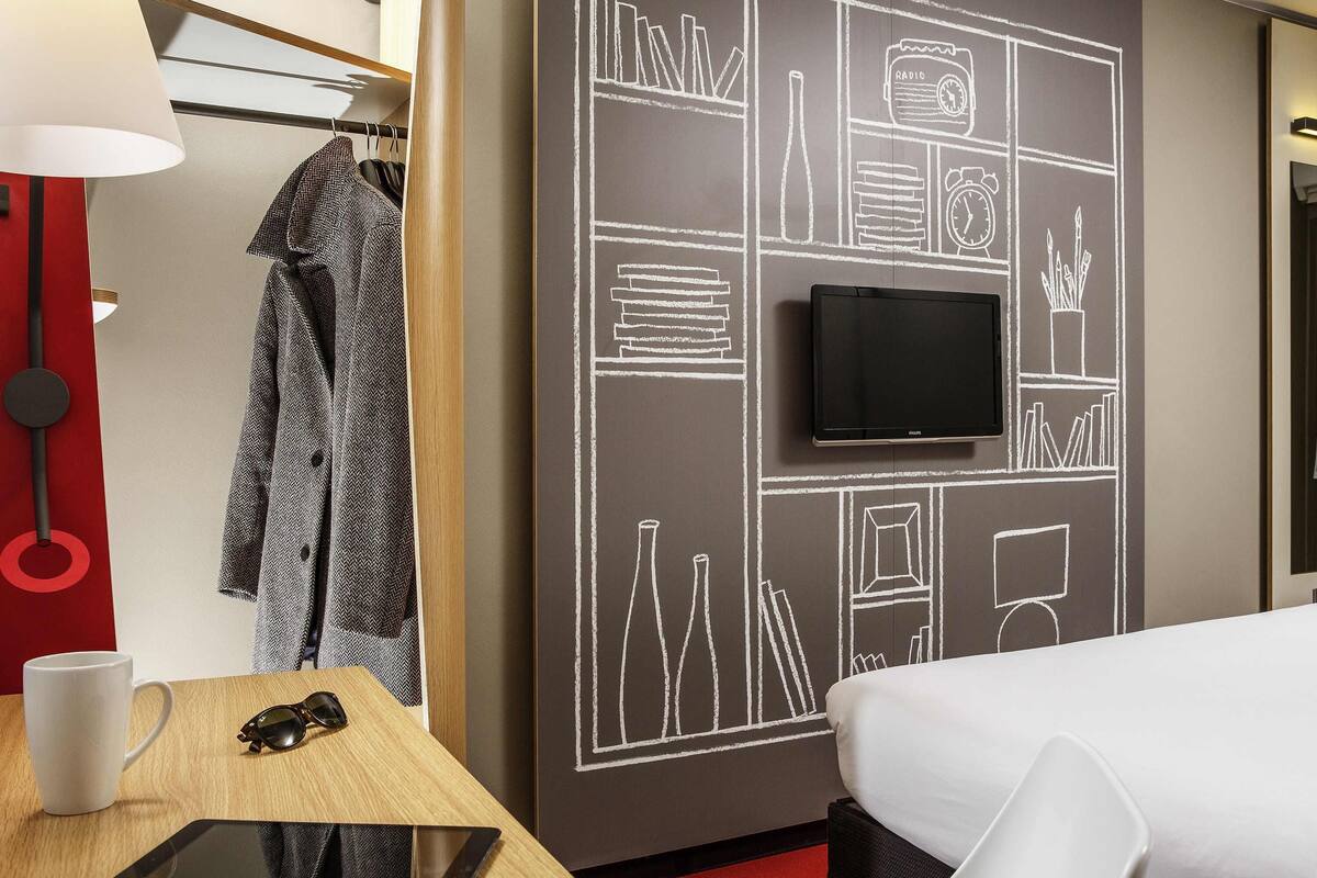 A hotel room with a bed, a desk with a tablet, a coffee cup, and a coat rack with a coat.