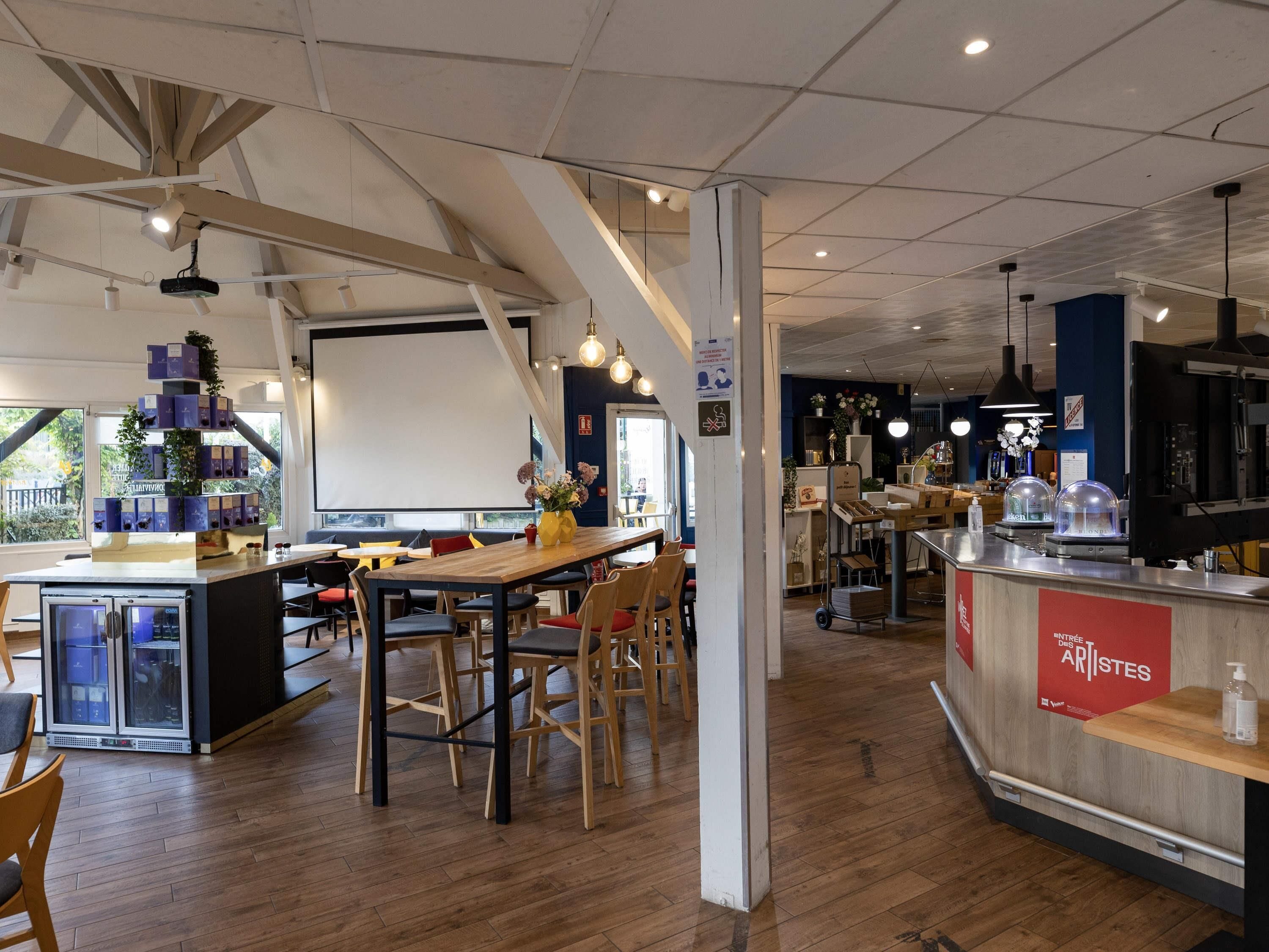 A modern cafe interior with wooden tables and chairs, a counter with a red sign, and a large window with a view of greenery outside.