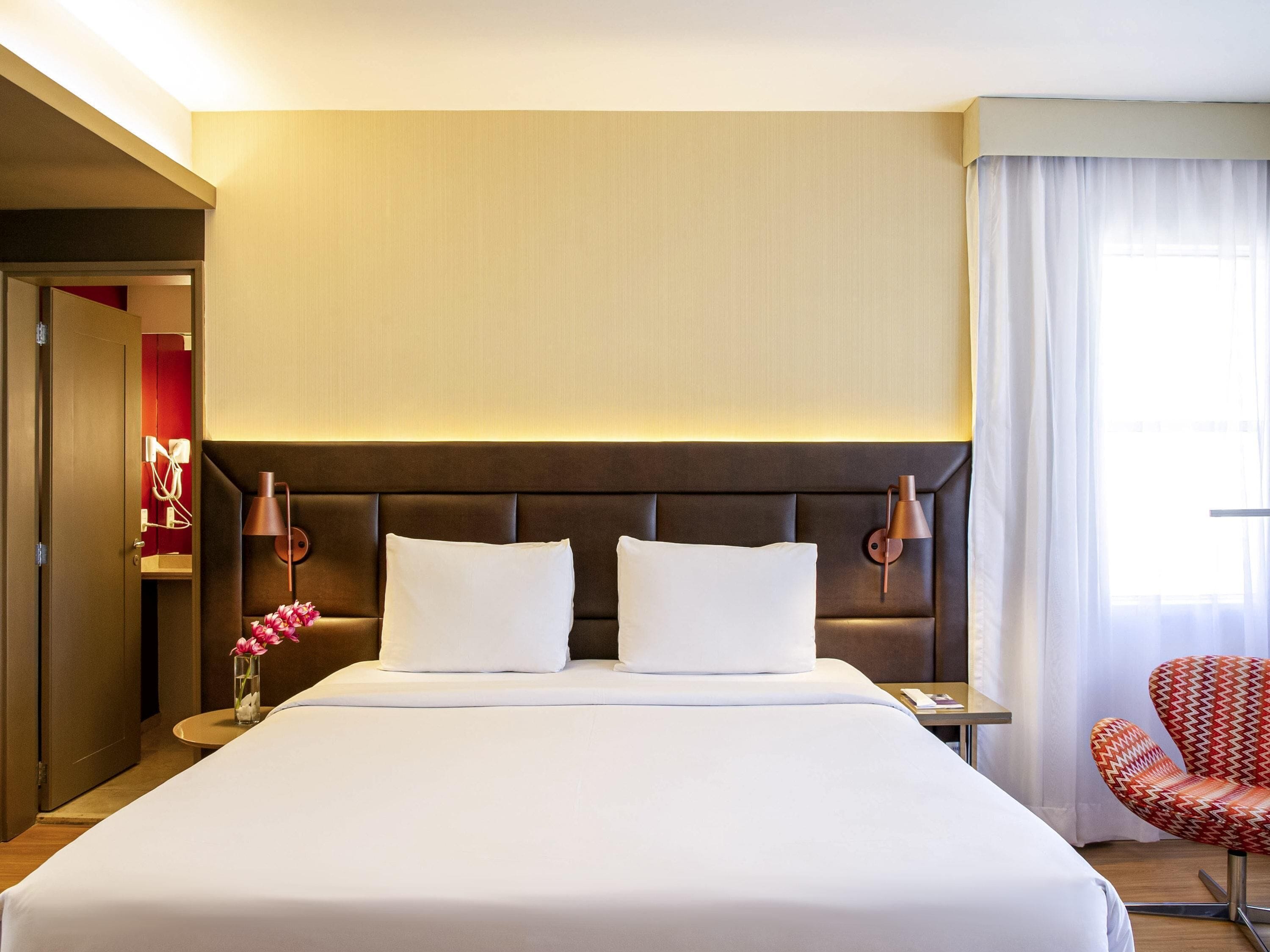 Een hotelkamer met een groot bed, een nachtkastje, een lamp, een stoel en een raam met gordijnen.