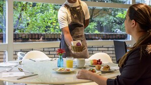 Une table mise pour le petit-déjeuner, avec une nappe pour deux personnes, comprenant des assiettes remplies de nourriture, une tasse de café et un panier de fruits.