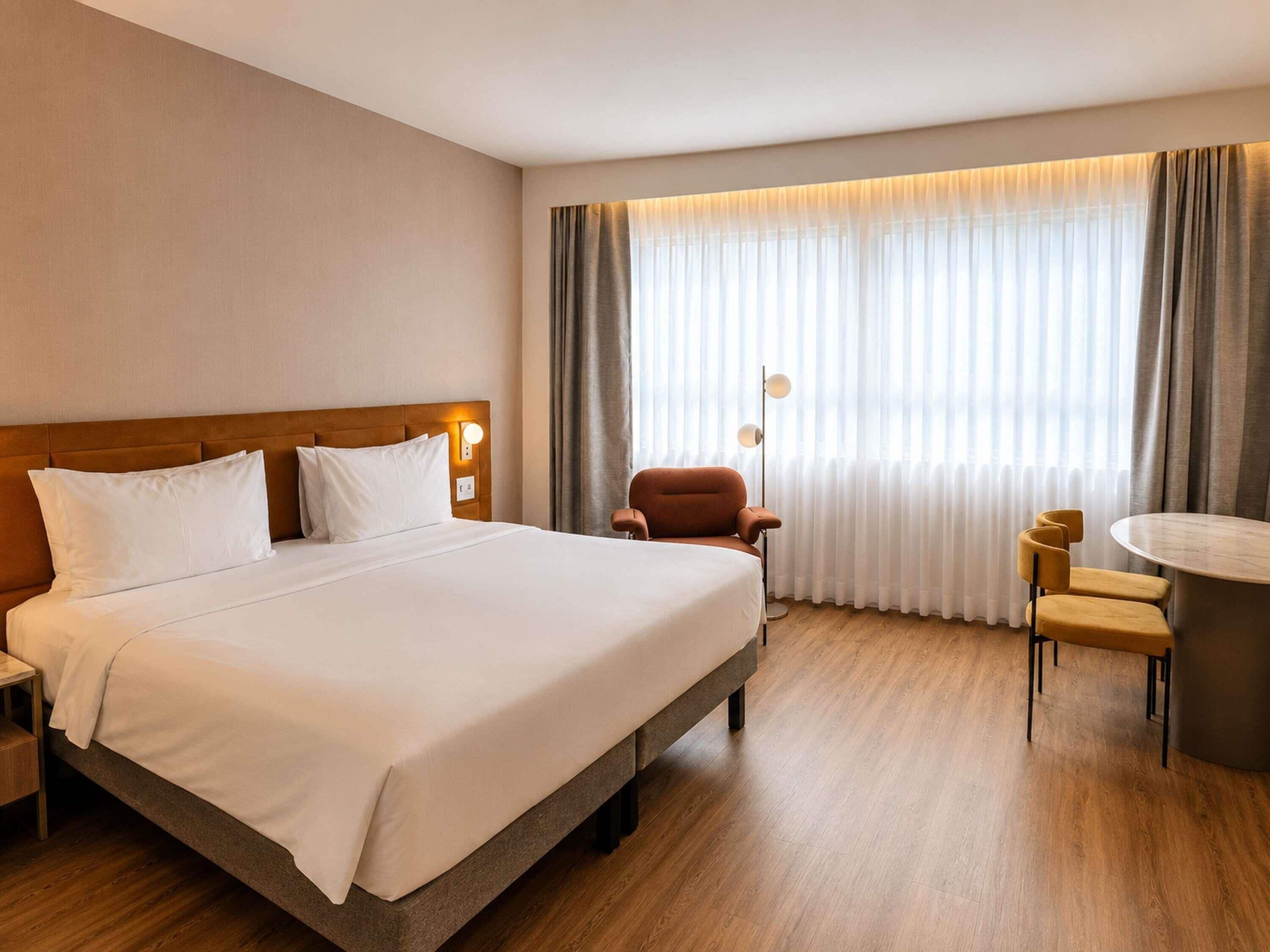 Una habitación de hotel con cama, una silla, un escritorio y una ventana con cortinas.