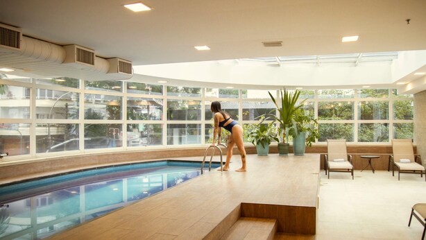 Une femme en maillot de bain, debout au bord d’une piscine, avec une vue sur la ville à travers de grandes fenêtres.
