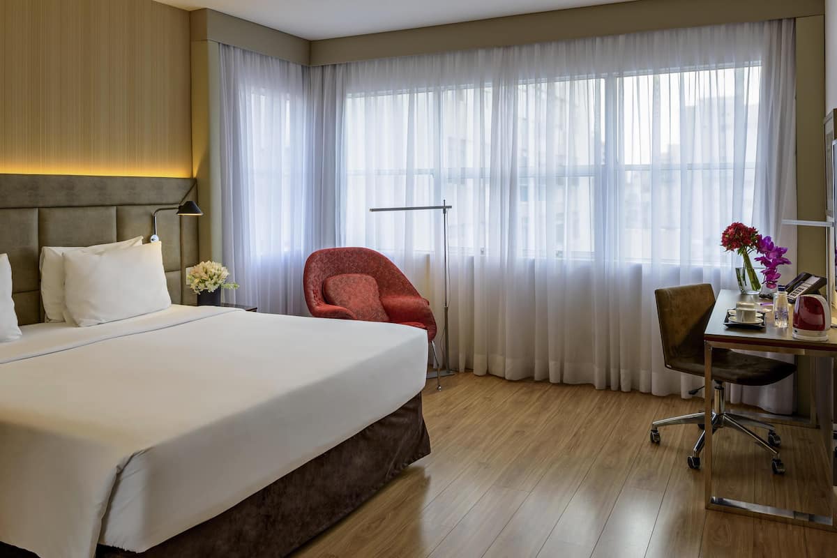 Ein Hotelzimmer mit einem großen Bett, einem Schreibtisch mit Stuhl, einem roten Sessel und einem großen Fenster mit transparenten Vorhängen.
