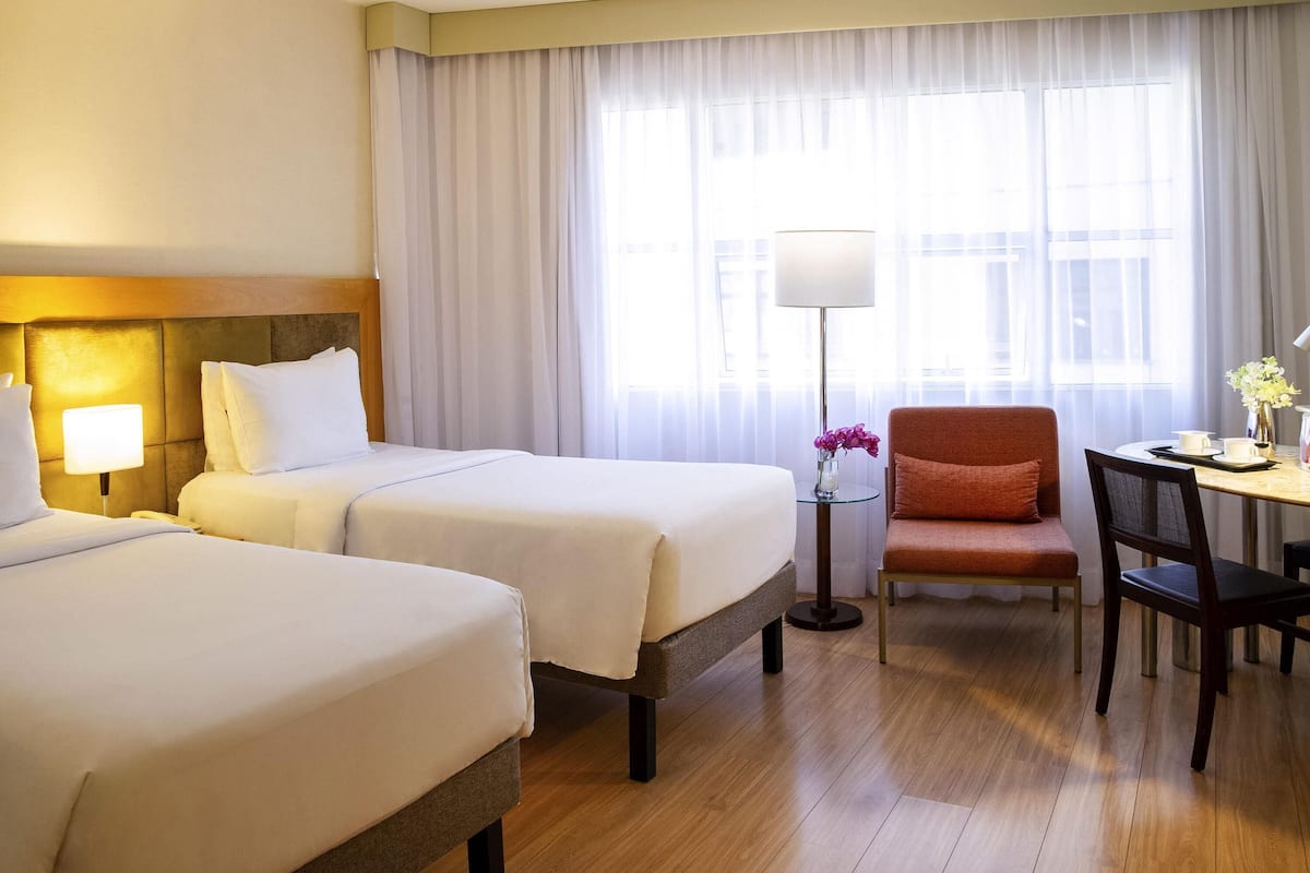 Ein Hotelzimmer mit zwei Betten, einem Schreibtisch mit Stuhl, einer Lampe und Blick durch große Fenster nach draußen.