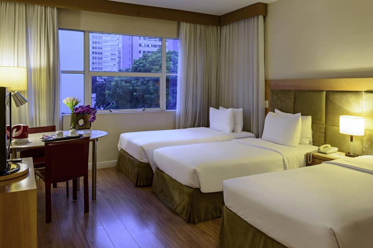 Ein Hotelzimmer mit zwei Betten, einem Schreibtisch mit Lampe, einem Sessel und einem Fenster mit Vorhängen.