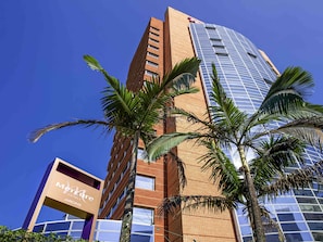 Hotel com fachada moderna em vidro e estrutura de tijolos, rodeado por palmeiras.