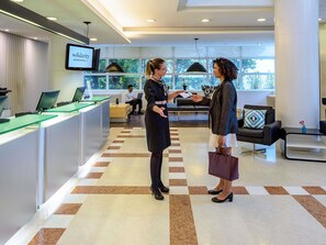 A reception area with a check-in counter, a television screen displaying the hotel name, and a seating area with a sofa and a coffee table. - Mercure Sao Paulo Jardins (São Paulo)