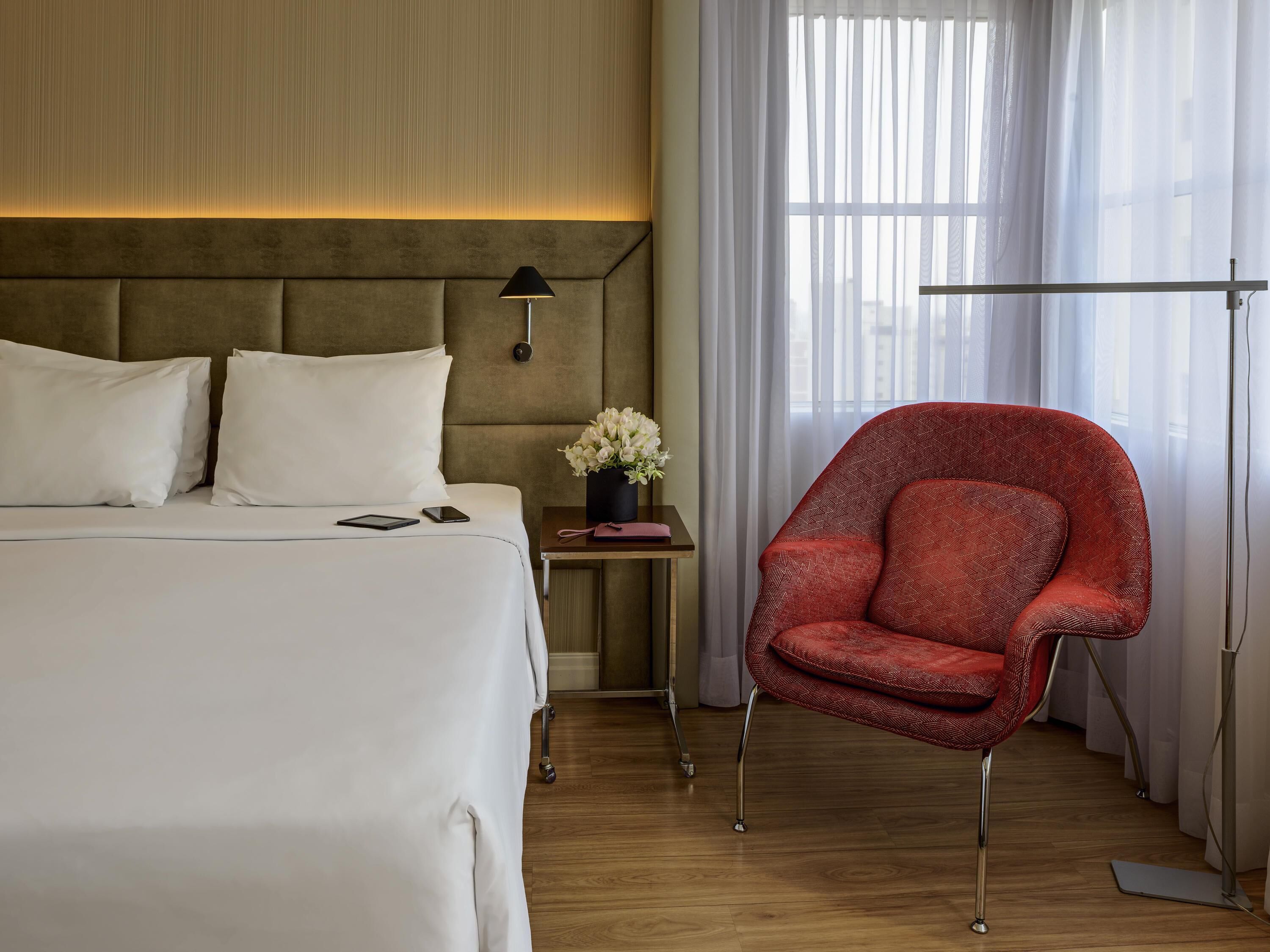 Une chambre d’hôtel avec un lit, une table de chevet, une lampe, une chaise et une fenêtre avec des rideaux.