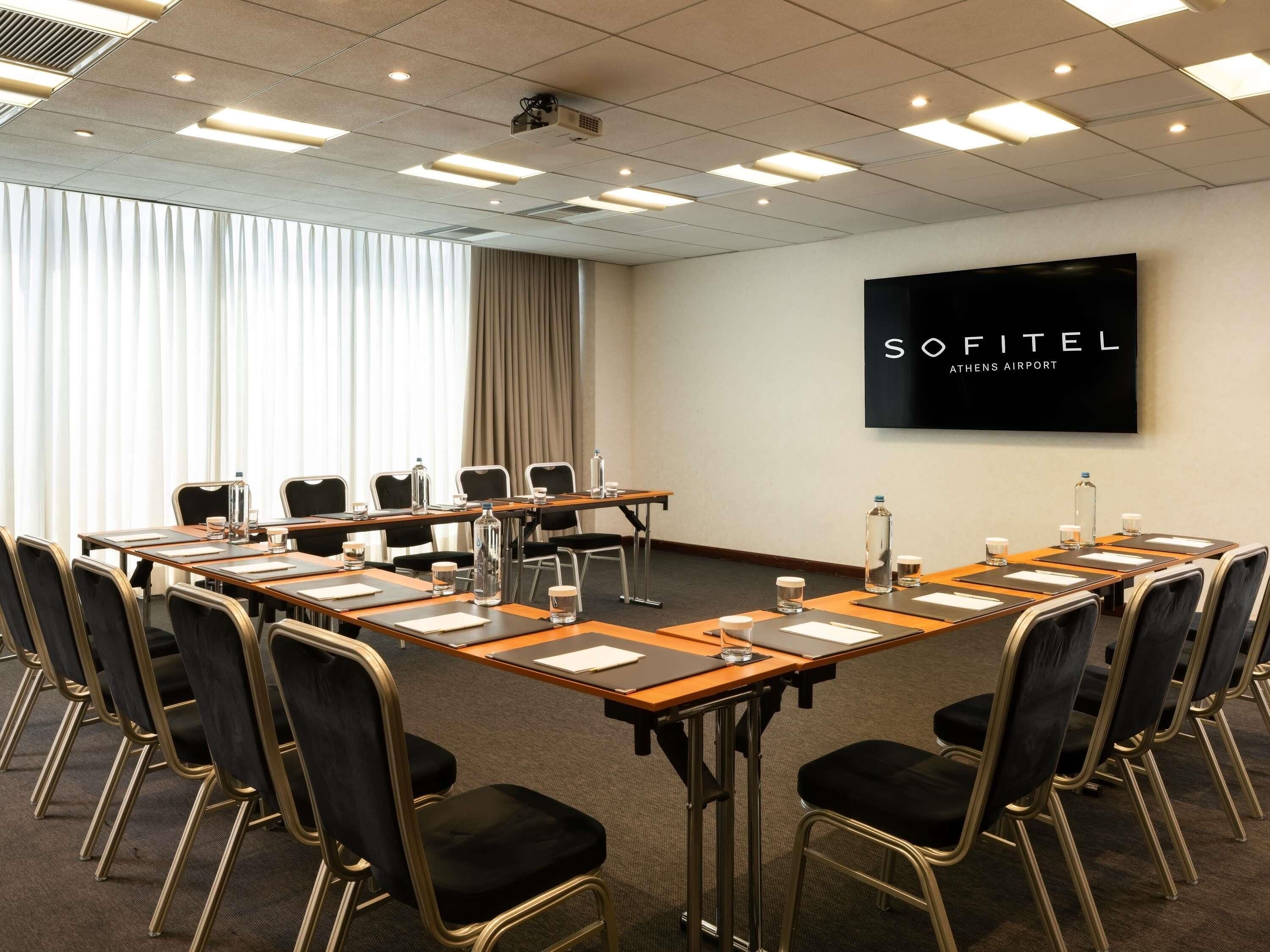 A conference room with a long table, chairs, and a projector screen displaying 'SOFITEL ATHENS AIRPORT'.