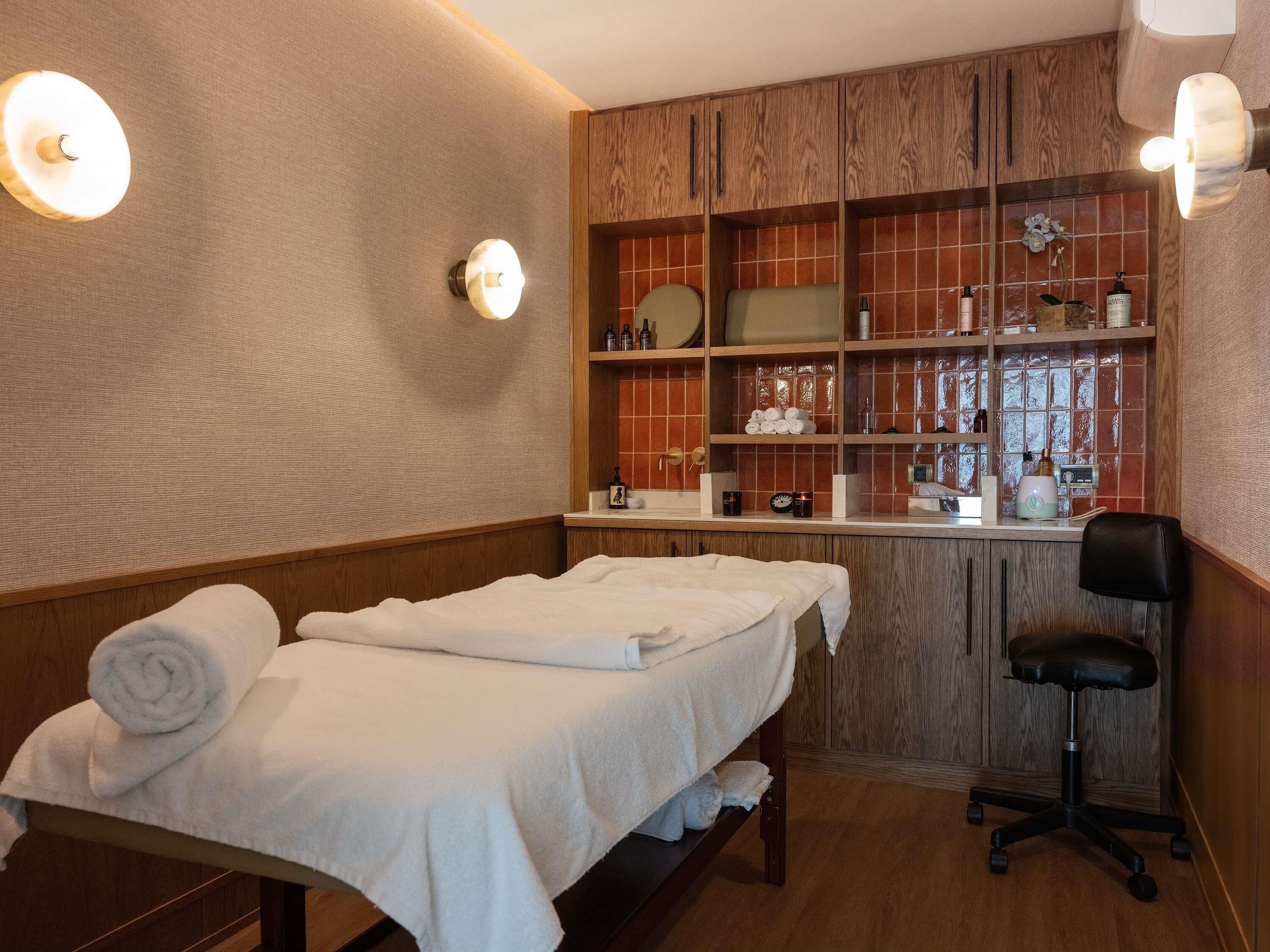 Um quarto de spa com mesa de massagem, uma cadeira, prateleiras com produtos de higiene pessoal e um espelho.