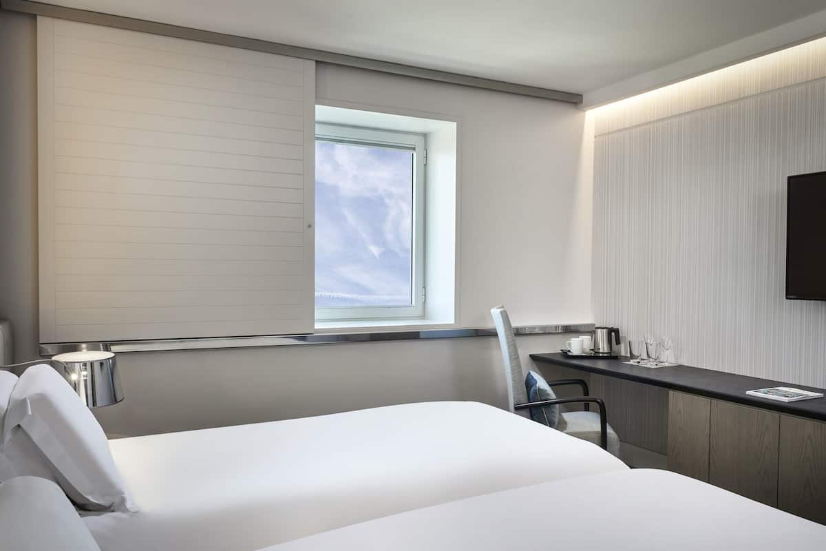 A modern hotel room with a bed, a desk, a chair, a window, and a flat-screen TV.