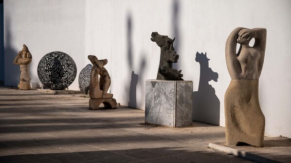 Una hilera de esculturas al aire libre frente a una pared blanca, con sombras proyectadas sobre el suelo.