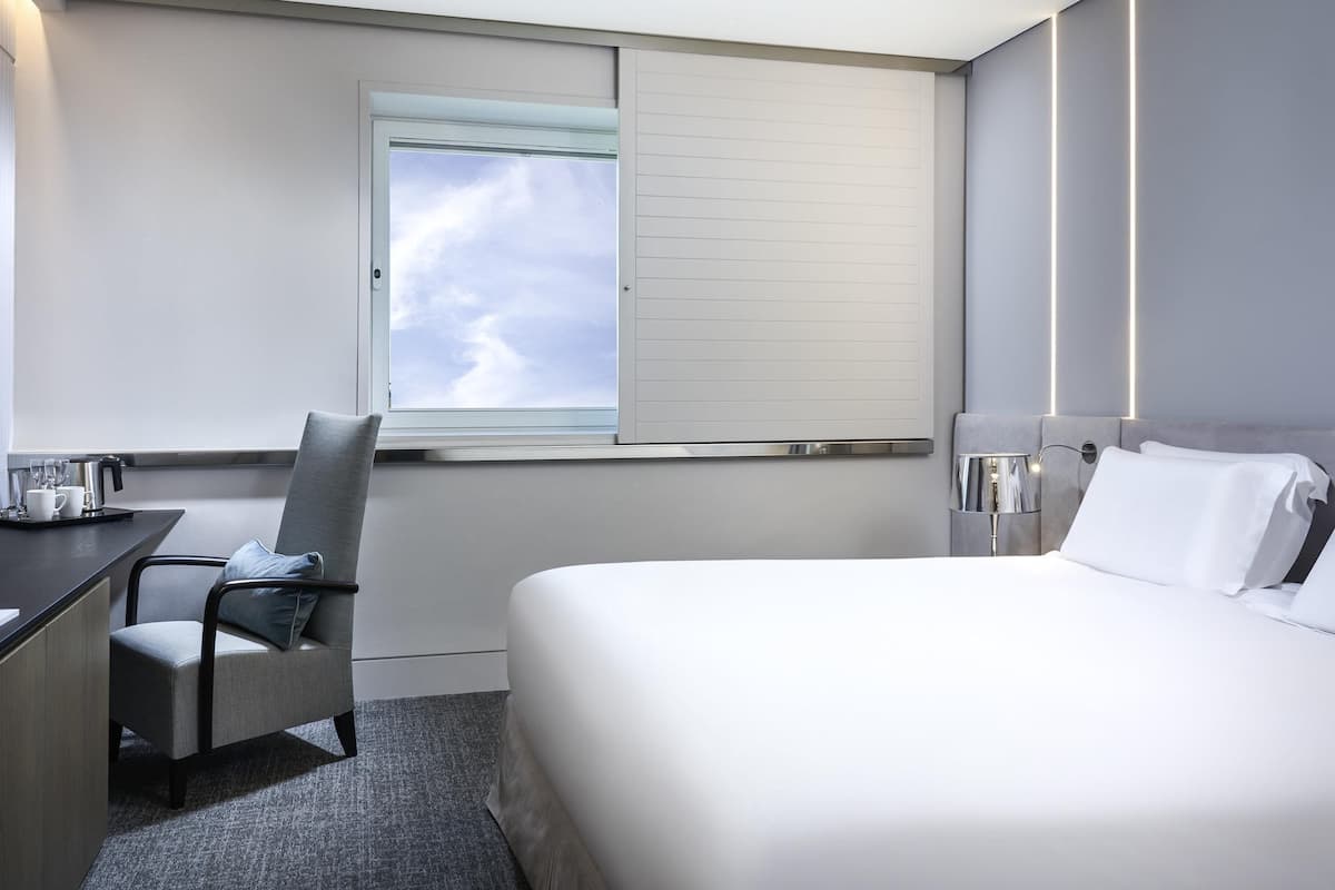 A hotel room with a bed, a desk, a chair, a window with a view of the sky, and a bedside lamp.