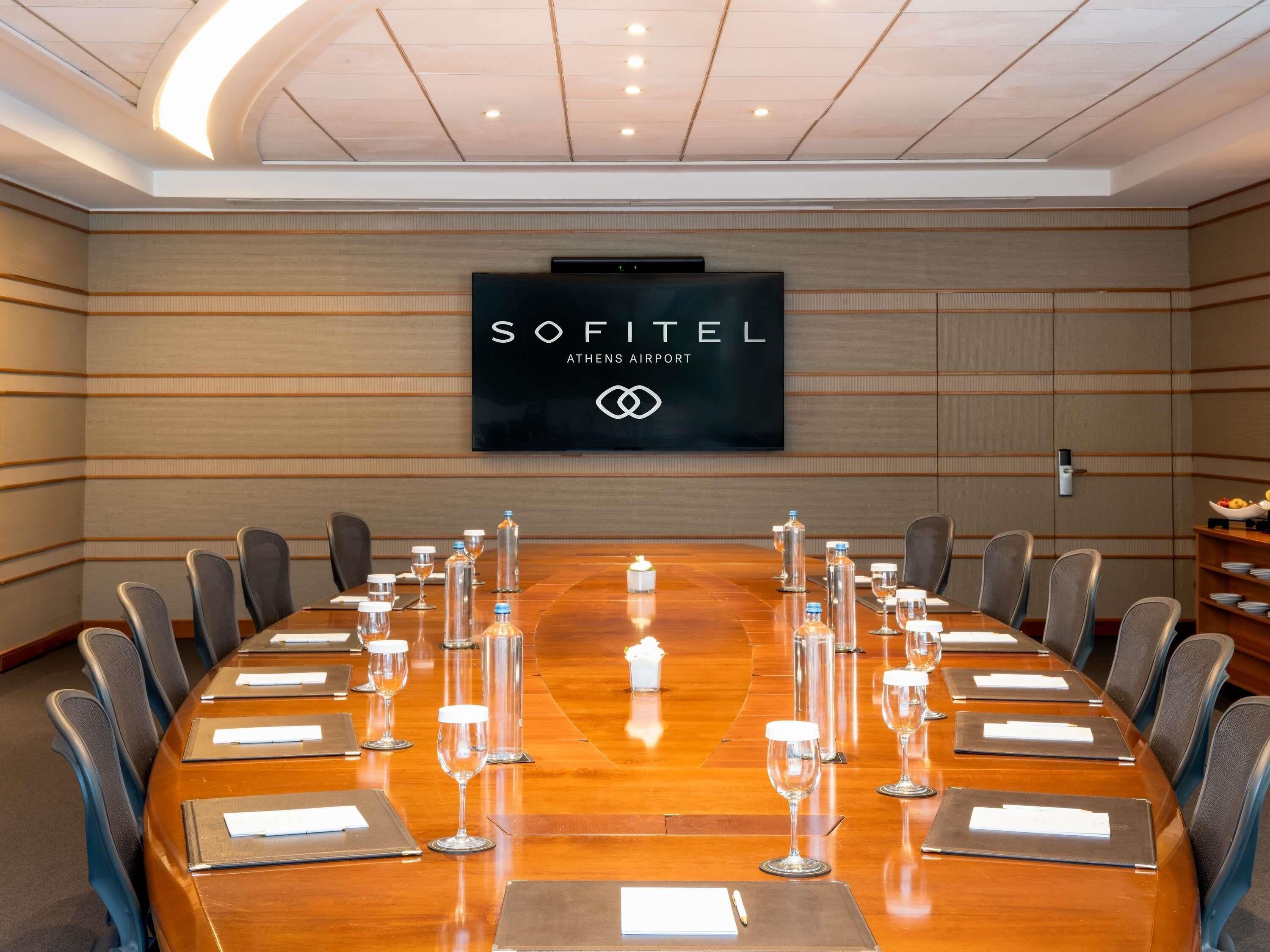 Un salón de conferencias con una larga mesa, sillas, placas con nombres y un cartel de Sofitel Athens Airport.