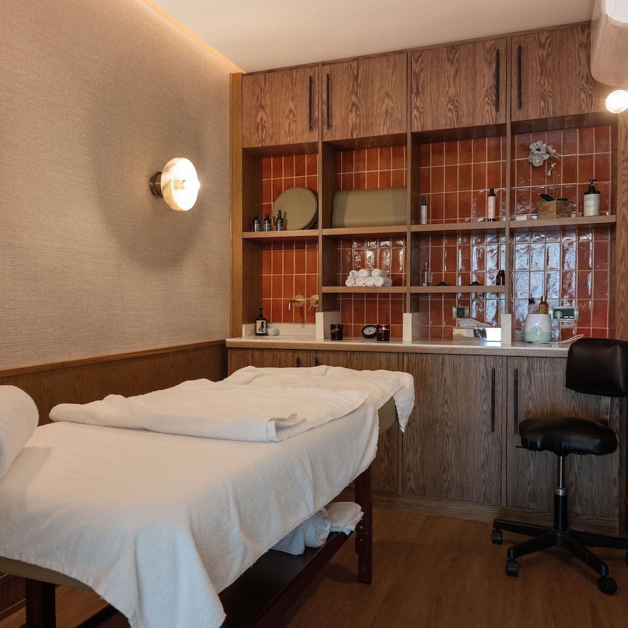 A spa room with a massage table, a chair, shelves with toiletries, and a mirror.