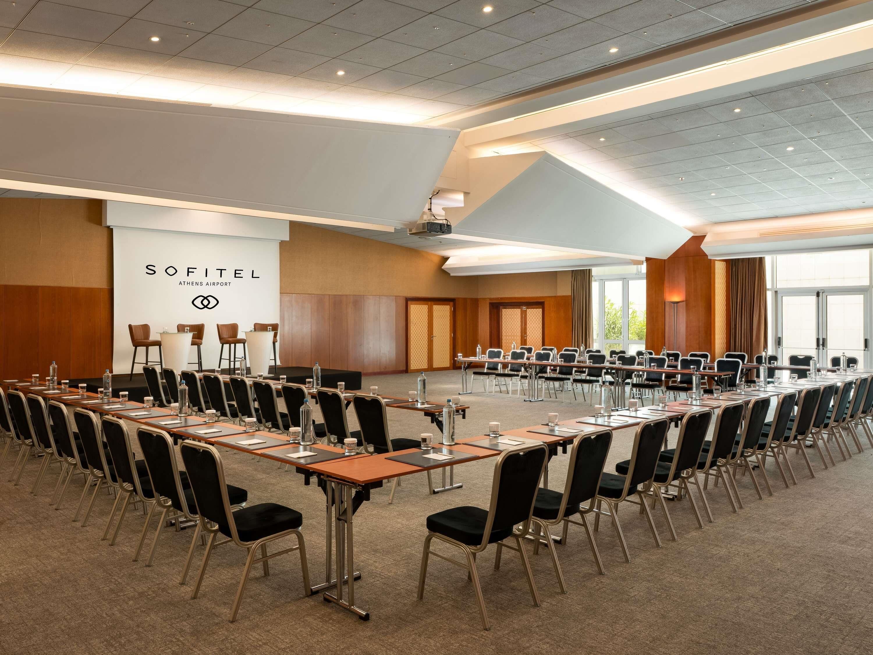 Ein Konferenzraum mit langem Tisch, Stühlen und einer Präsentationsleinwand im Sofitel Athens Airport.