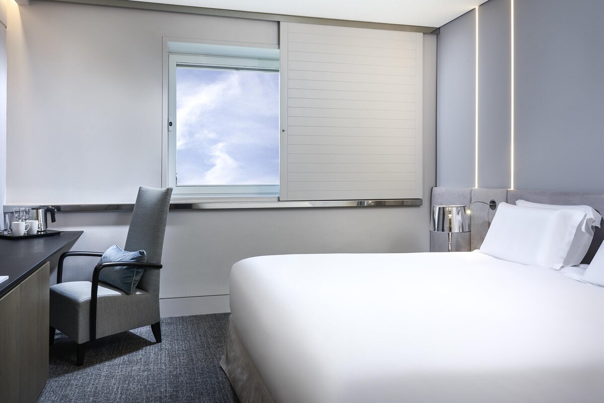 Ein Hotelzimmer mit Bett, Schreibtisch, Stuhl, einem Fenster mit Blick auf den Himmel und einer Nachttischlampe.