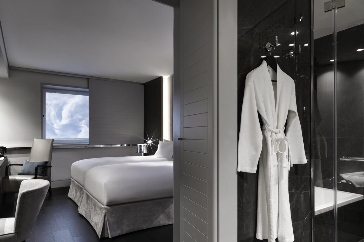 A modern hotel room with a large bed, a seating area with a chair and a table, a robe hanging on a rack, and a glass shower enclosure.