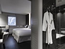 Una habitación de hotel moderna con una cama grande, una zona de estar con una silla y una mesa, un albornoz colgado en un perchero y un box de ducha de vidrio.