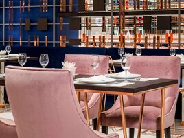 Interior de un restaurante con sillas tapizadas en rosa, mesas de madera oscura y una pared azul con lámparas verticales.