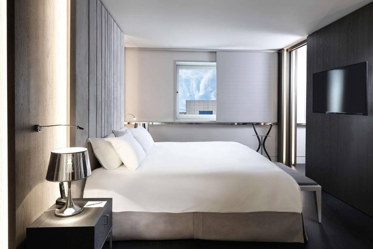 A modern hotel room with a large bed, a bedside table with a lamp, a TV mounted on the wall, and a window with a view of the sky.