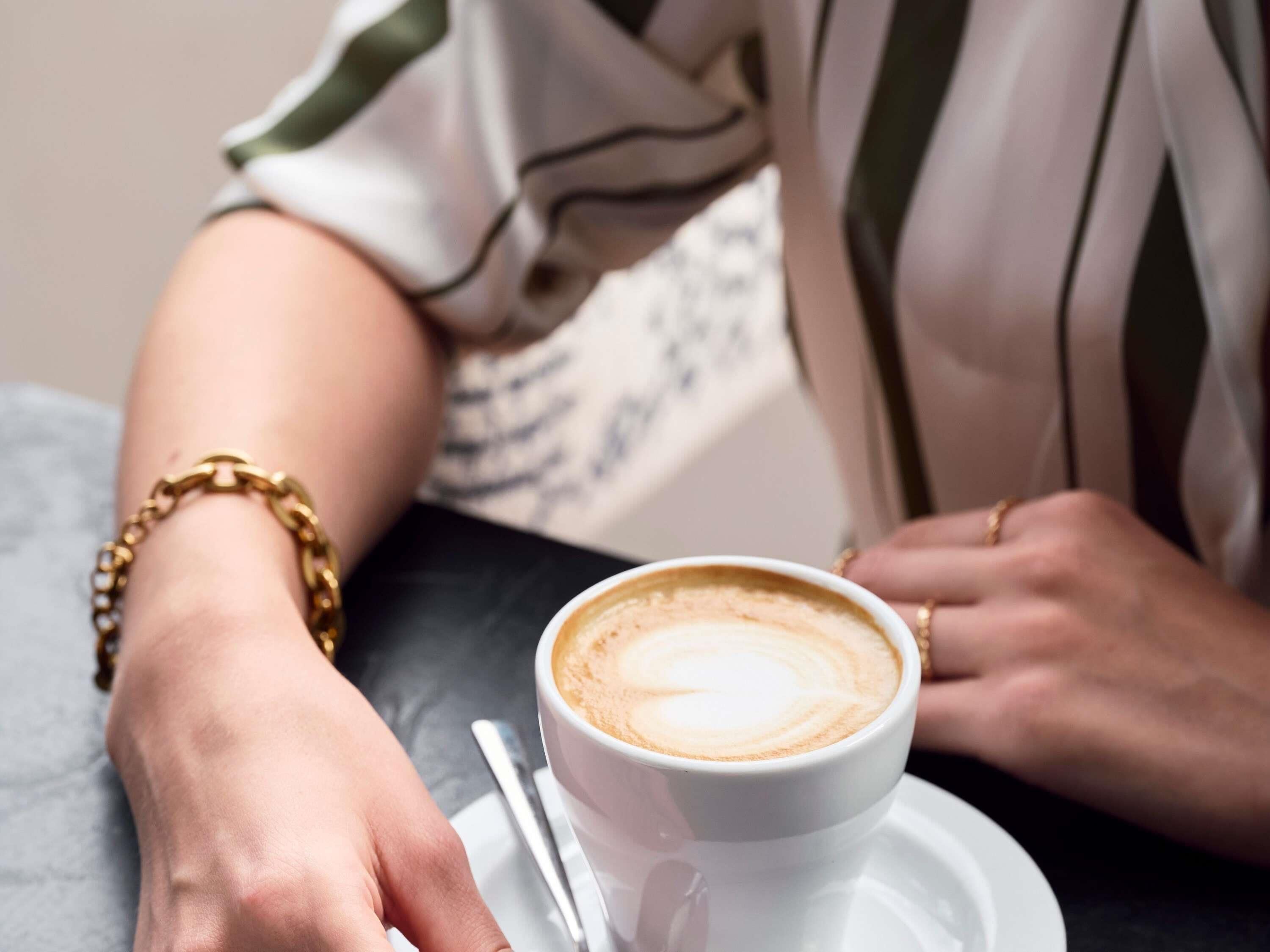 Eine Kaffeetasse auf einem Untertasse mit einem Löffel, eine Hand mit einem goldenen Armband und einem gestreiften Hemd.