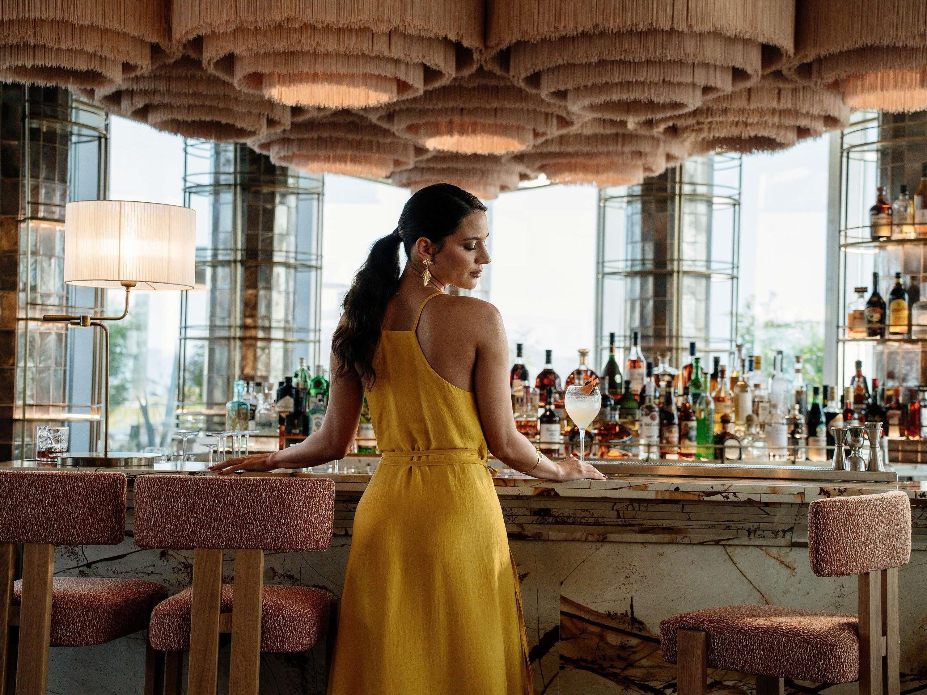 A woman in a yellow dress at a bar with a view of the city through large windows.