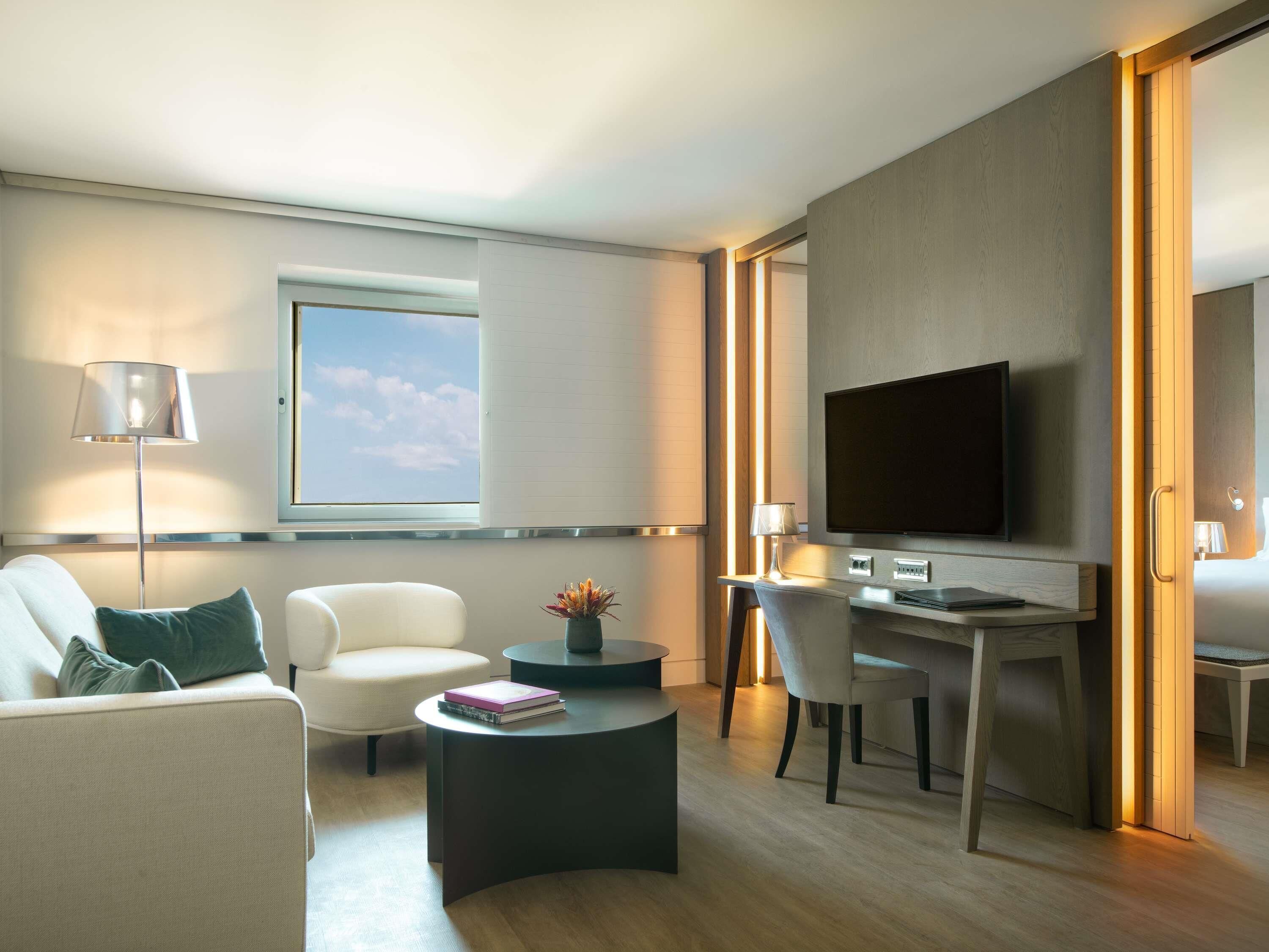 Una habitación de hotel moderna con sofá, escritorio, silla, televisor y ventana con persianas.
