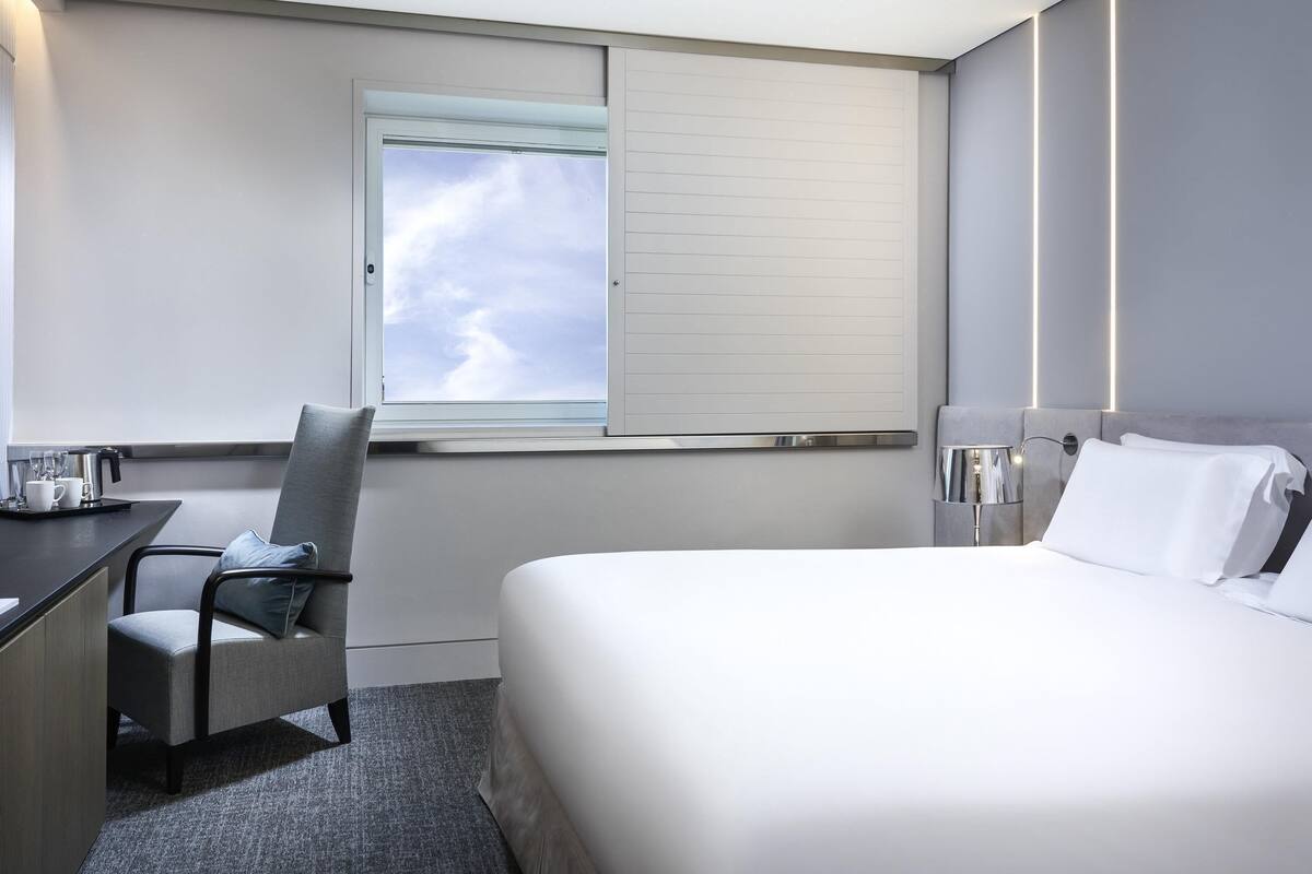 A hotel room with a bed, a desk, a chair, a window with a view of the sky, and a nightstand with a lamp.