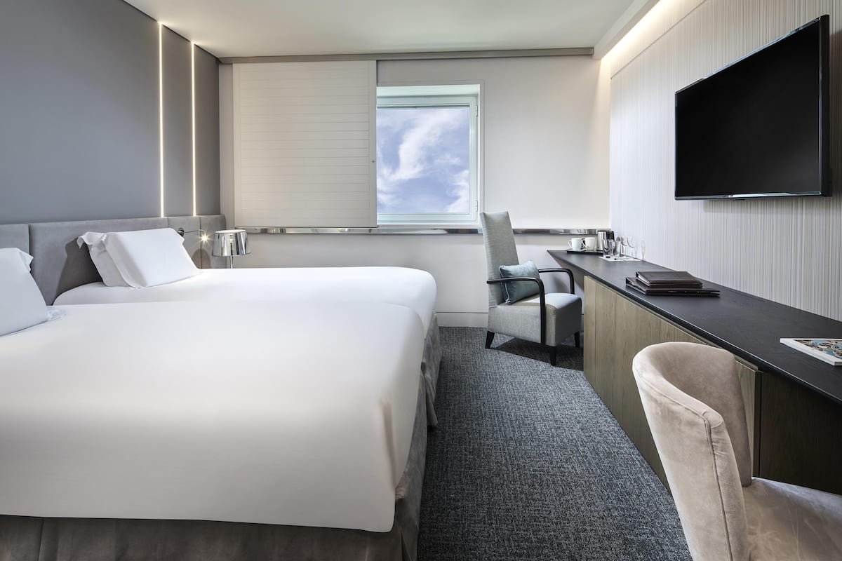 A modern hotel room with a large bed, a desk, a chair, a flat-screen TV, and a window with a view of the sky.