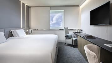 A modern hotel room with a large bed, a desk, a chair, a flat-screen TV, and a window with a view of the sky.