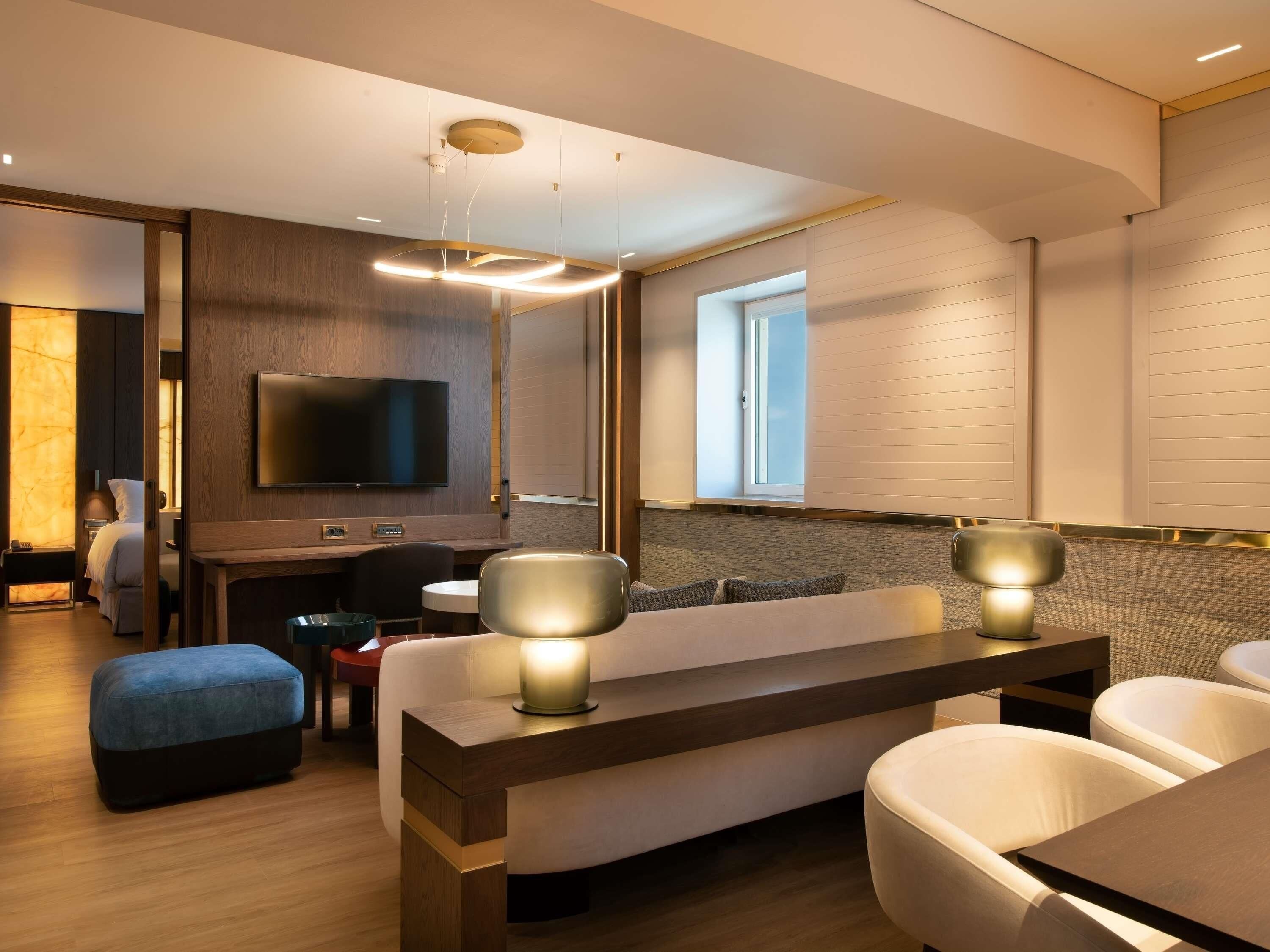 Ein modernes Hotelzimmer mit Holzboden, einer Couch, einem Esstisch, einem Fernseher und einem Fenster mit Jalousien.