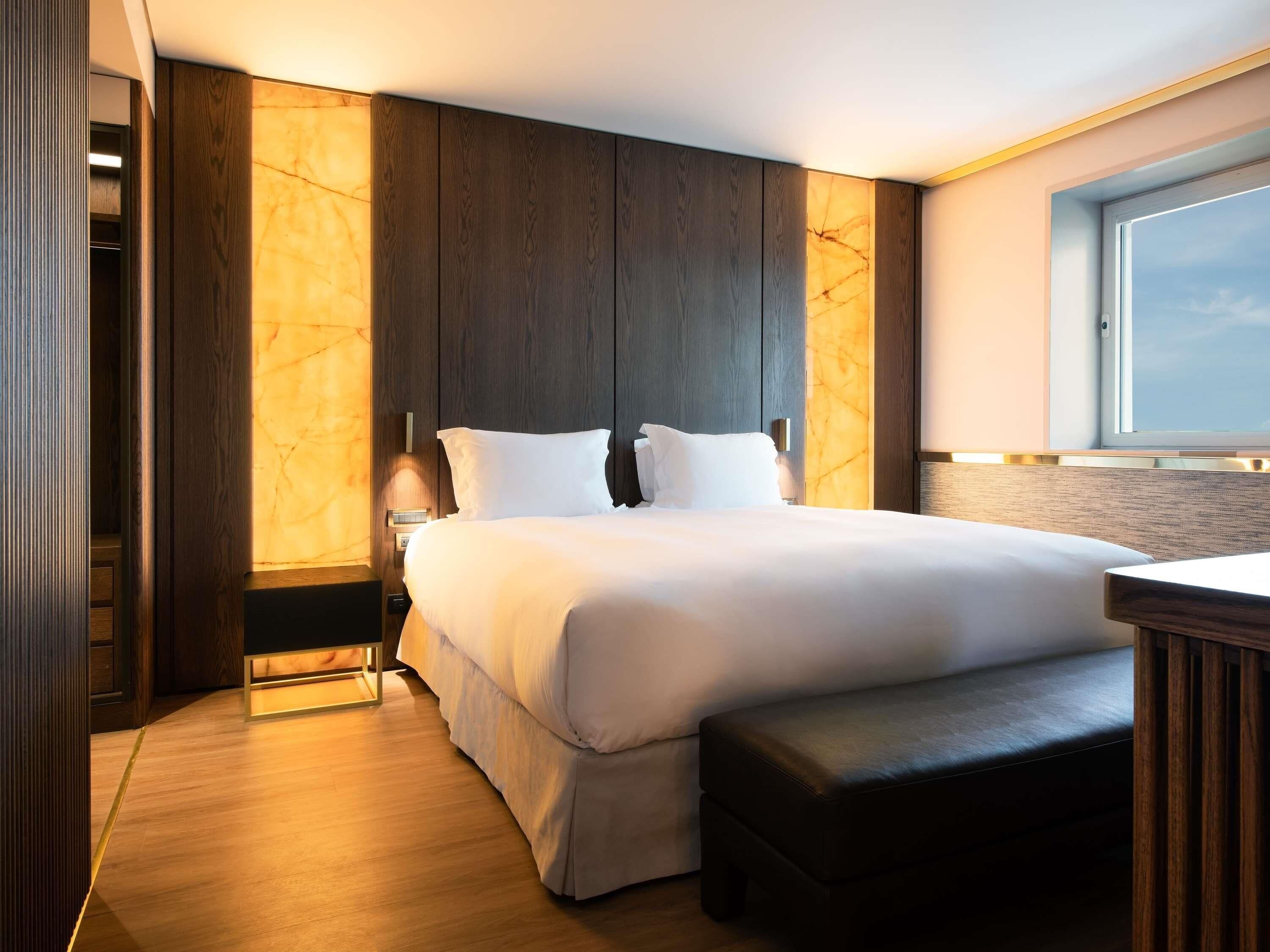 Una habitación de hotel moderna con una cama grande, cabecero de madera, una mesita de noche y una ventana.