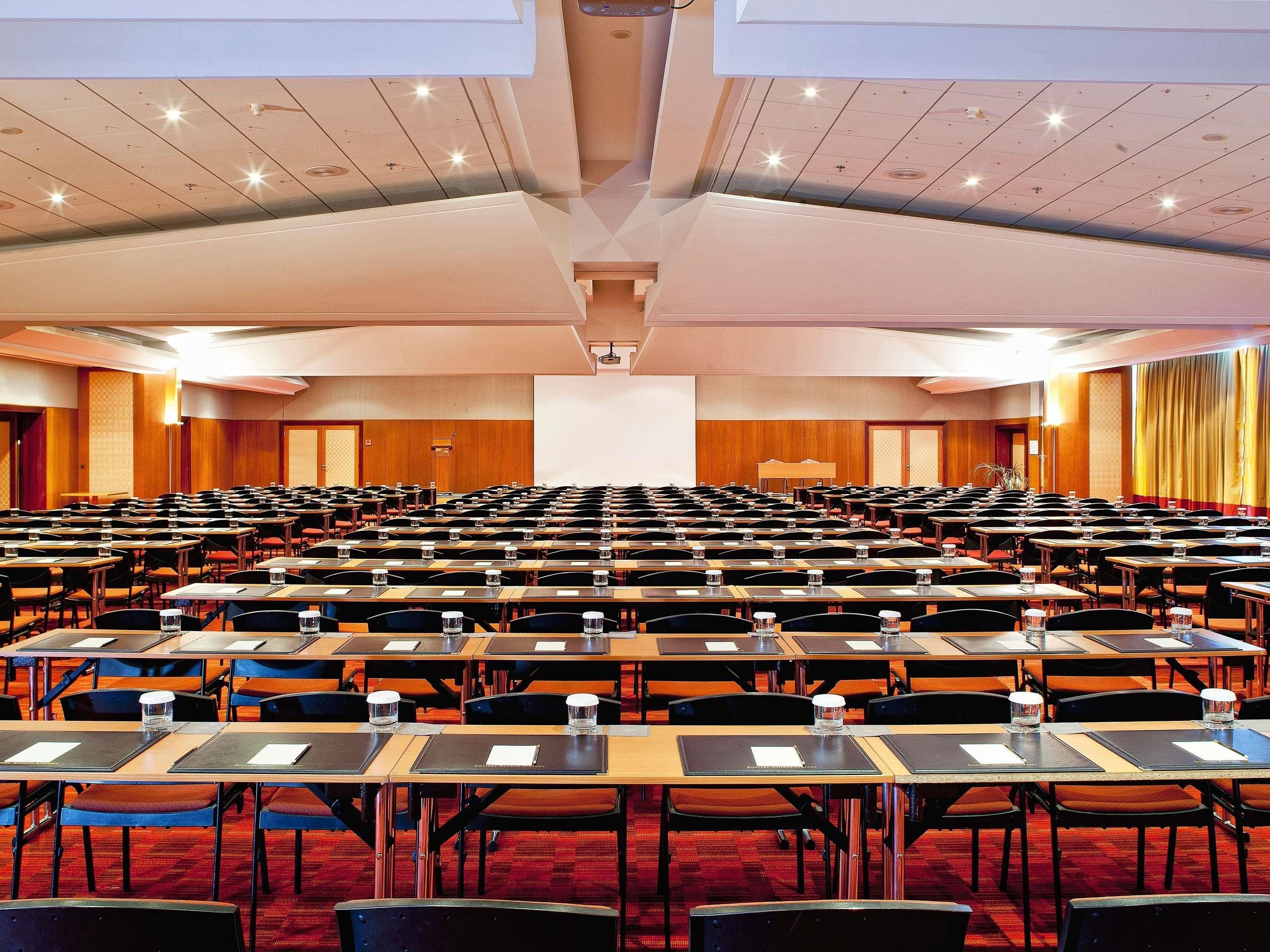 A conference room with rows of tables and chairs, a projector screen, and a stage area.
