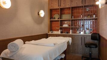 A spa room with a massage table, a chair, shelves with toiletries, and a mirror.