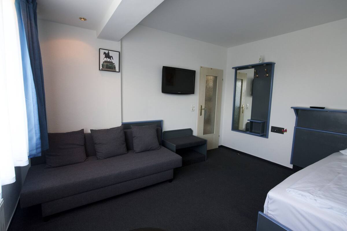 Habitación de hotel con un sofá gris, una cama, un televisor y un espejo.