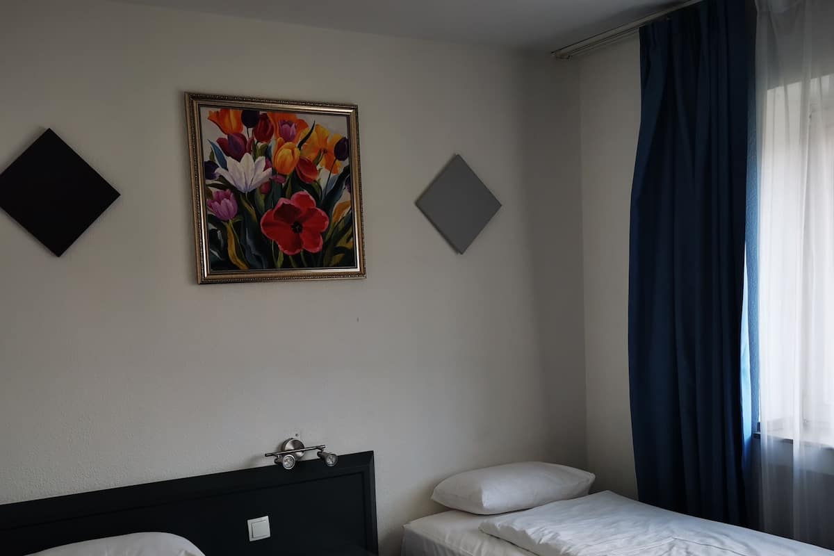 Habitación de hotel con dos camas individuales, un cuadro de flores en la pared y una ventana con cortinas azules.