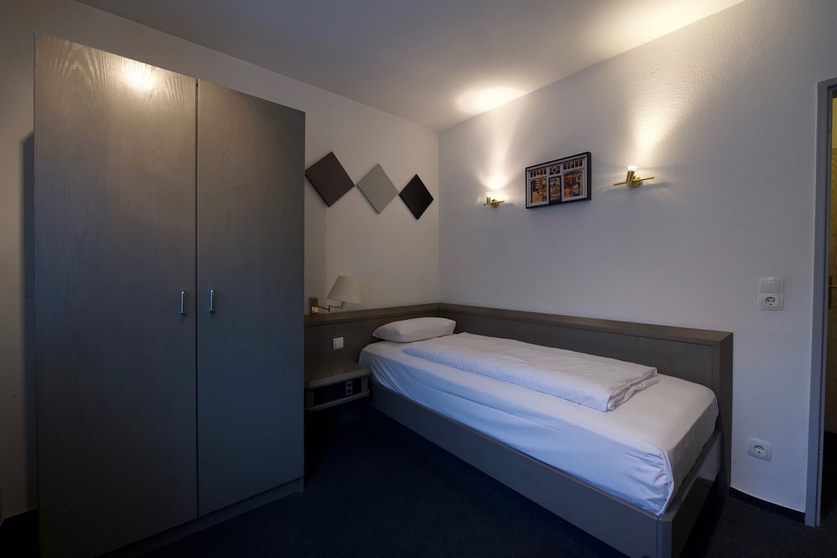 Una habitación de hotel con una cama individual, un armario, una mesita de noche y un cuadro colgado en la pared.
