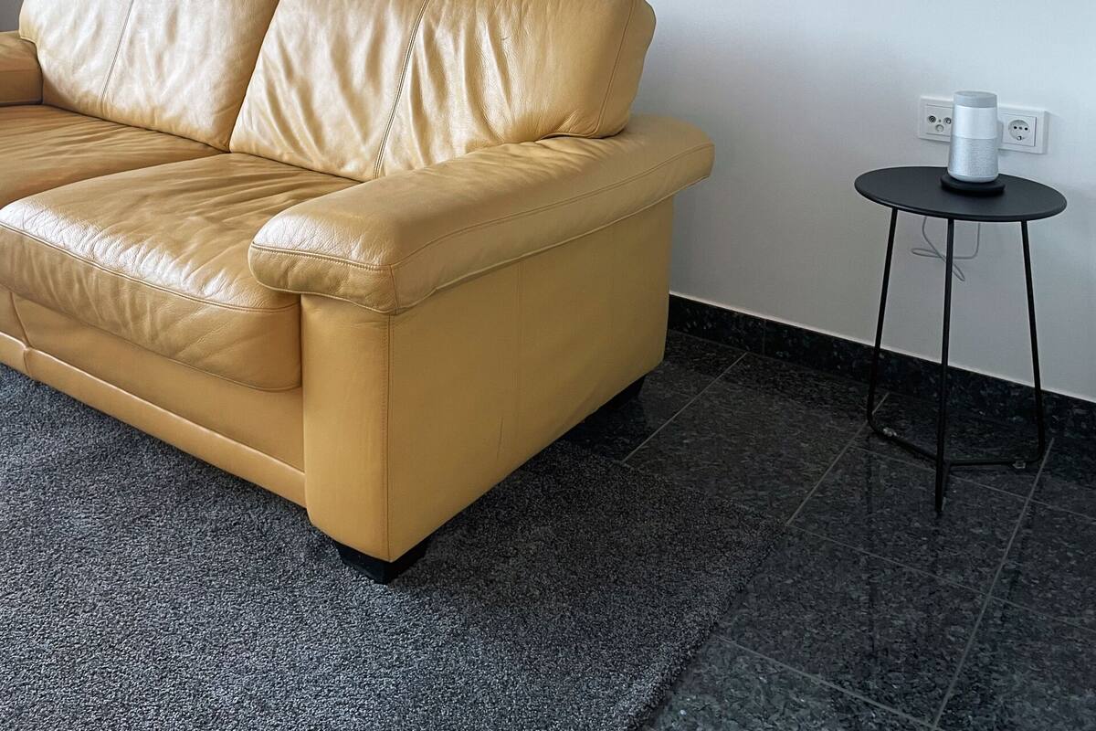 A room with a beige leather sofa, a framed artwork on the wall, a small table with a lamp, and a power outlet.