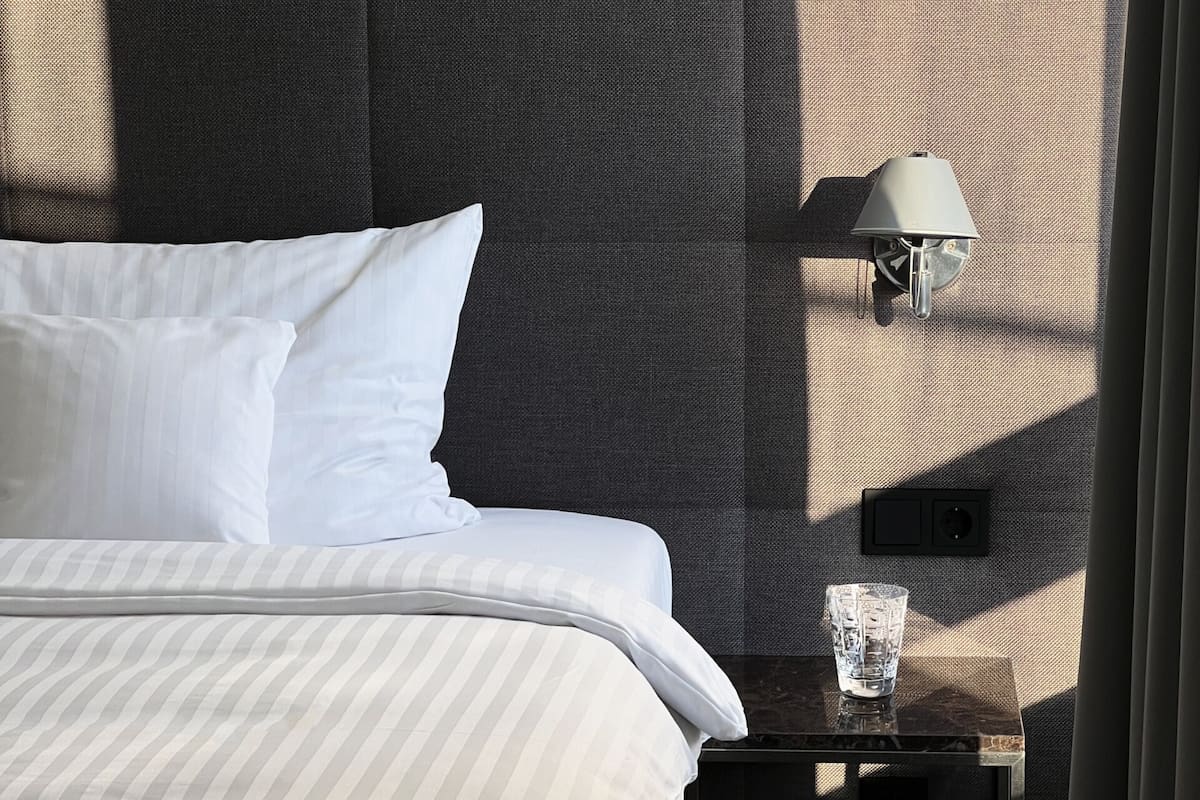 A neatly made bed with white linens, a wall-mounted lamp, and a small table with a glass.