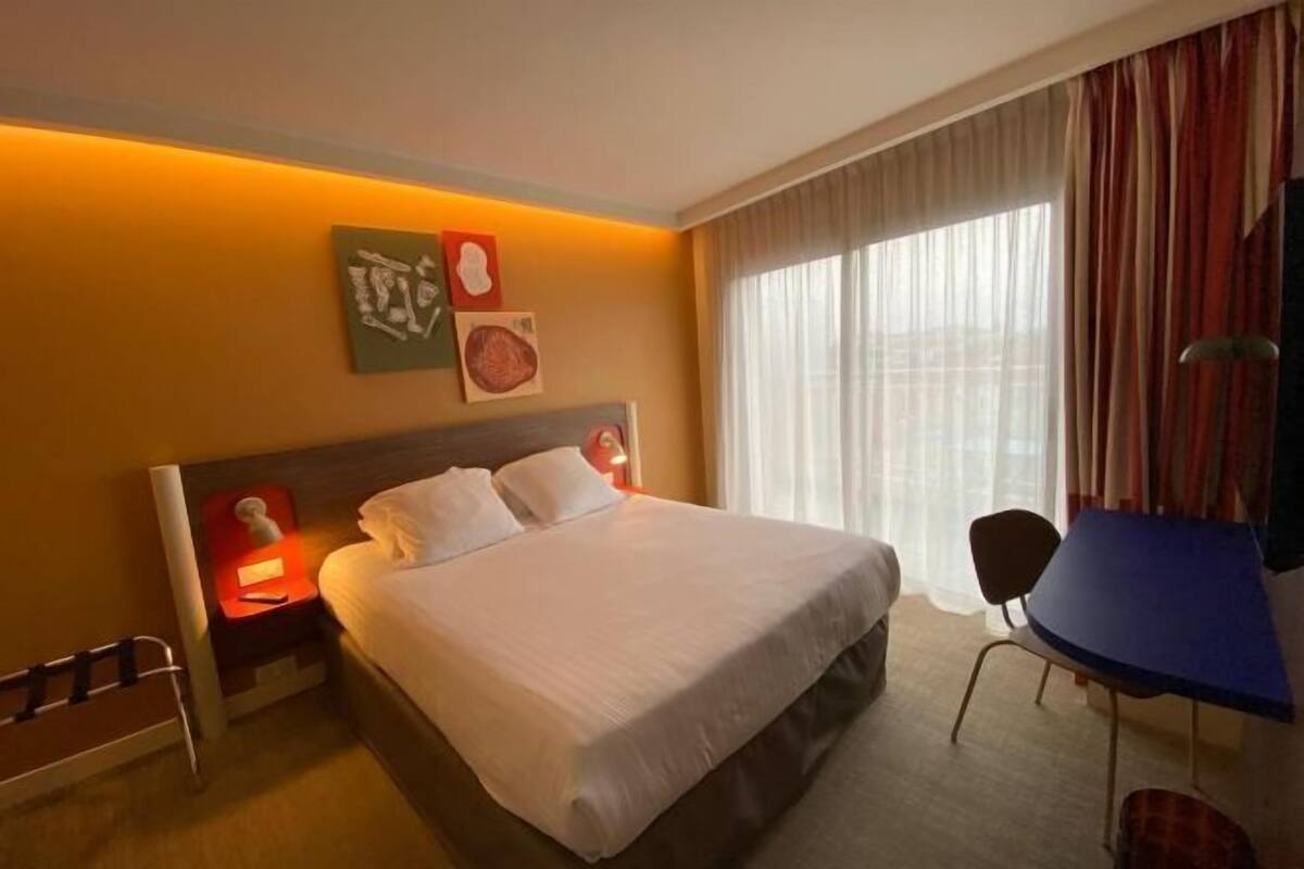 Habitación de hotel con cama, mesita de noche, lámpara, silla y vistas al exterior a través de la ventana.